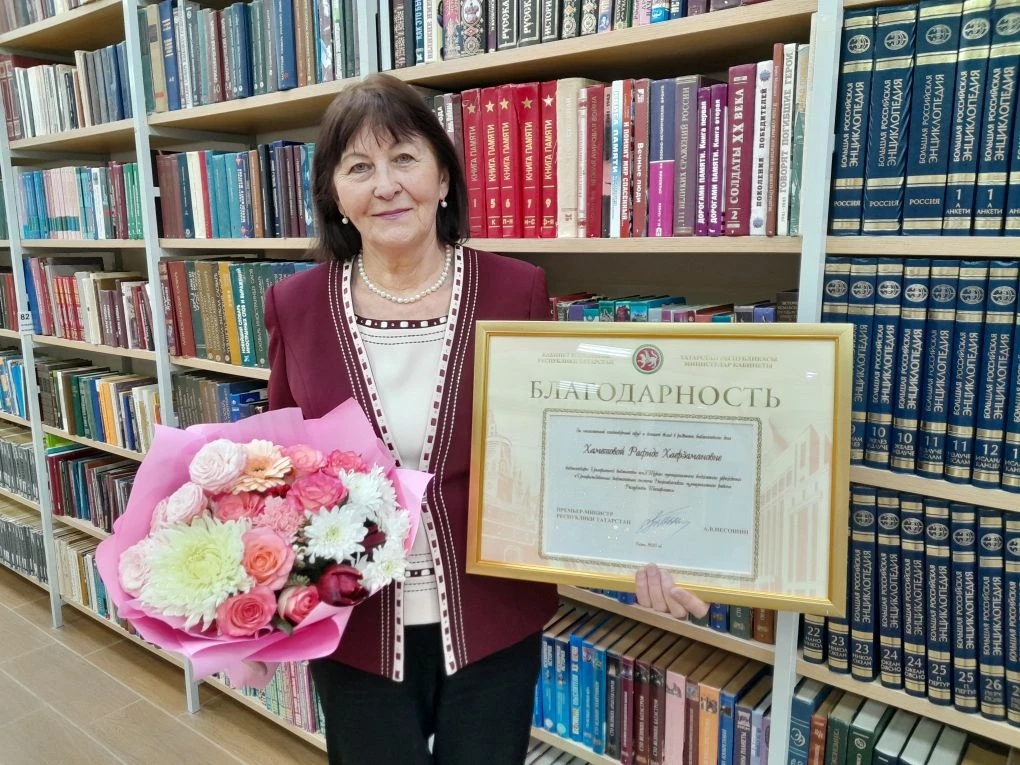 Хаметова Р.Х. стоит с букетом цветов и благодарственным письмом на фоне шкафов с книгами